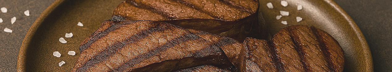 Grilled beef heart slices arranged in a fan shape on a rustic ceramic plate, with coarse sea salt scattered around on a dark brown textured surface.